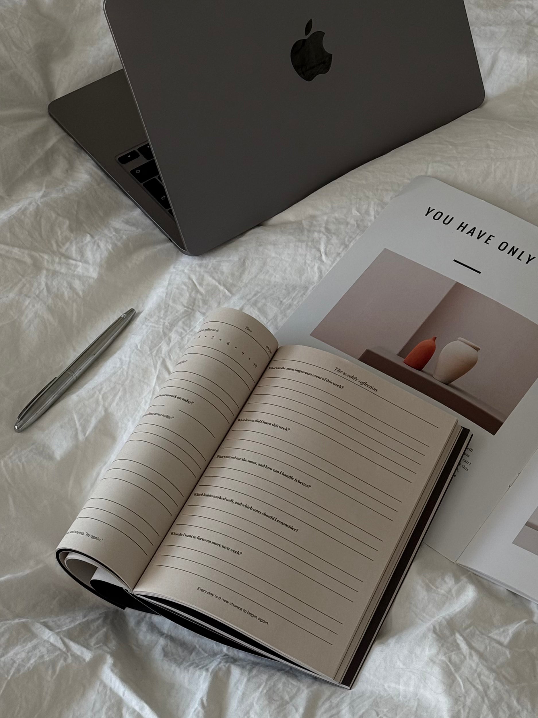 An open lined journal or notebook lies on white bedding alongside a closed gray laptop, a silver pen, and a printed card that reads 'YOU HAVE ONLY'.