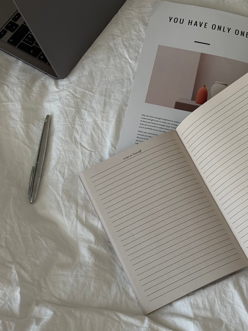 An open lined notebook lies on white wrinkled bedding next to a silver pen, with part of a laptop and an open magazine visible in a styled flat-lay arrangement.