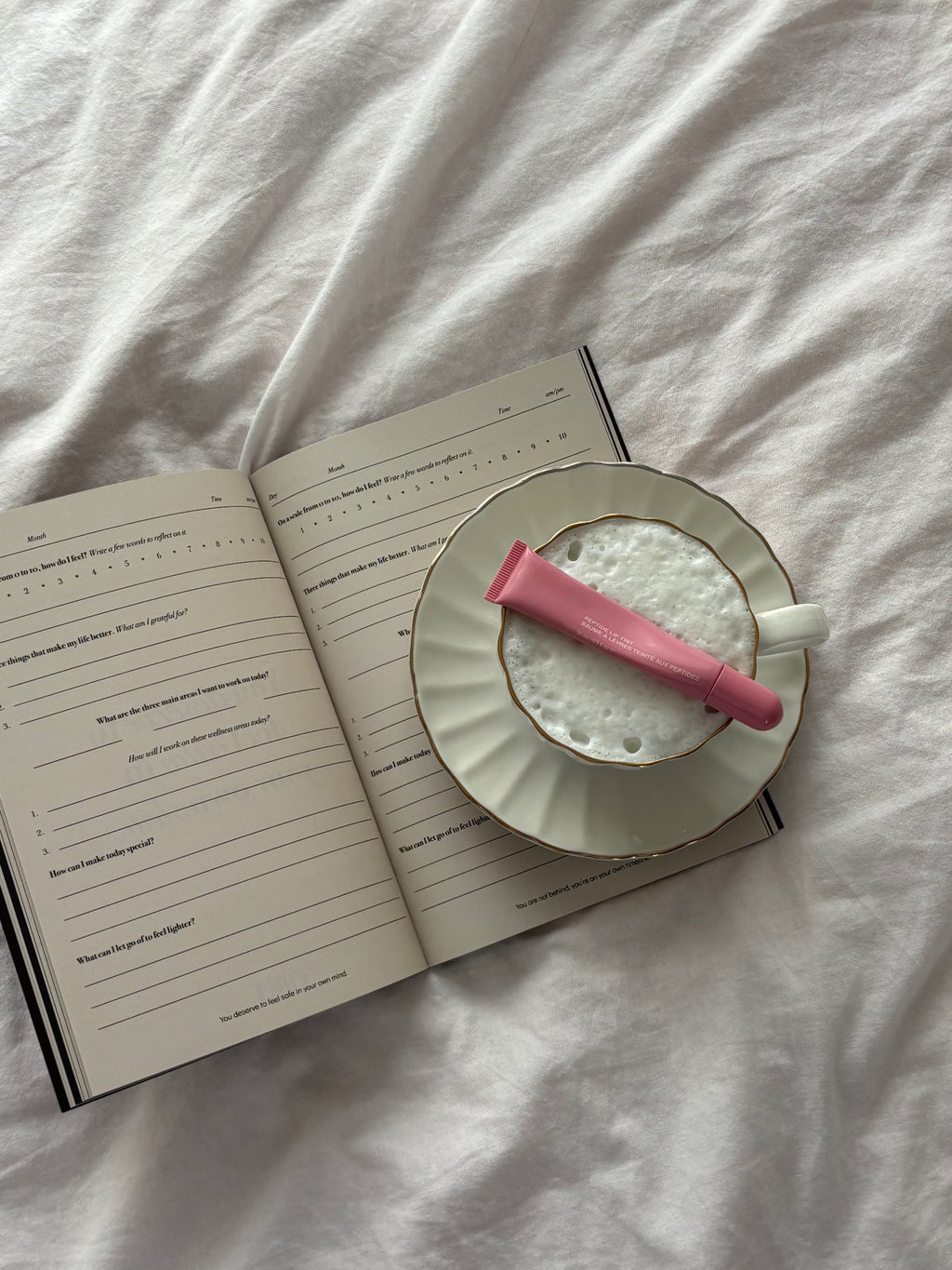 A pink lip balm tube rests on a white ceramic cup filled with white foam, placed on an open lined notebook over a rumpled white fabric background.