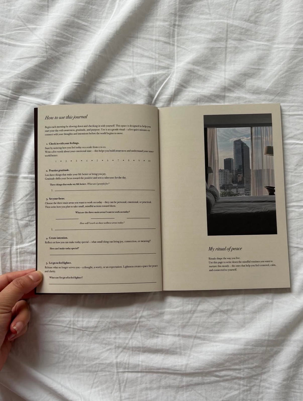 An open journal or notebook spread laid on white fabric, with printed instructions on the left page and a black-and-white cityscape photograph on the right page. A hand is holding the left edge of the journal.