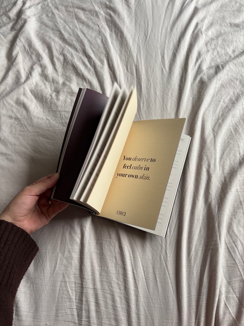 A hand holding an open book with cream-colored pages and a dark cover, set against a background of rumpled light gray fabric.