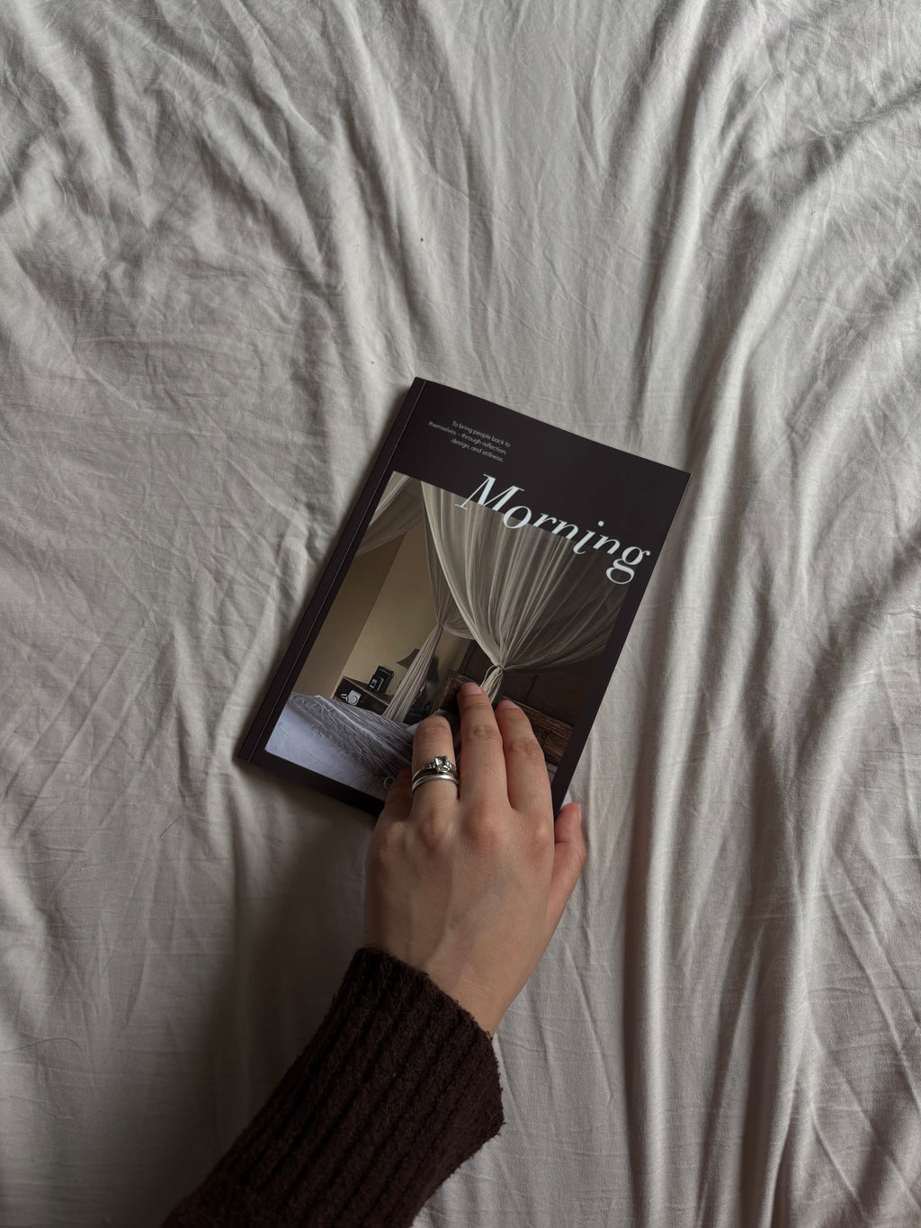 A hand wearing a dark brown ribbed sweater and a silver ring holds a book titled 'Morning' with a black cover, placed on a bed with rumpled white sheets.