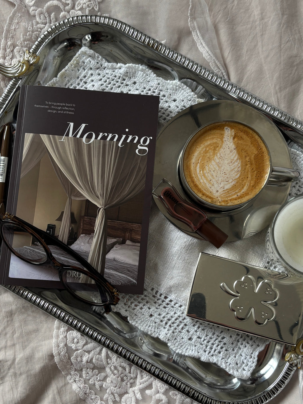 A styled flat-lay featuring a silver tray with a silver coffee cup of latte macchiato, a book titled 'Morning', eyeglasses, a small silver box, and a candle on a white lace doily over beige fabric.