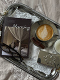 A styled flat-lay featuring a silver tray with a silver coffee cup of latte macchiato, a book titled 'Morning', eyeglasses, a small silver box, and a candle on a white lace doily over beige fabric.
