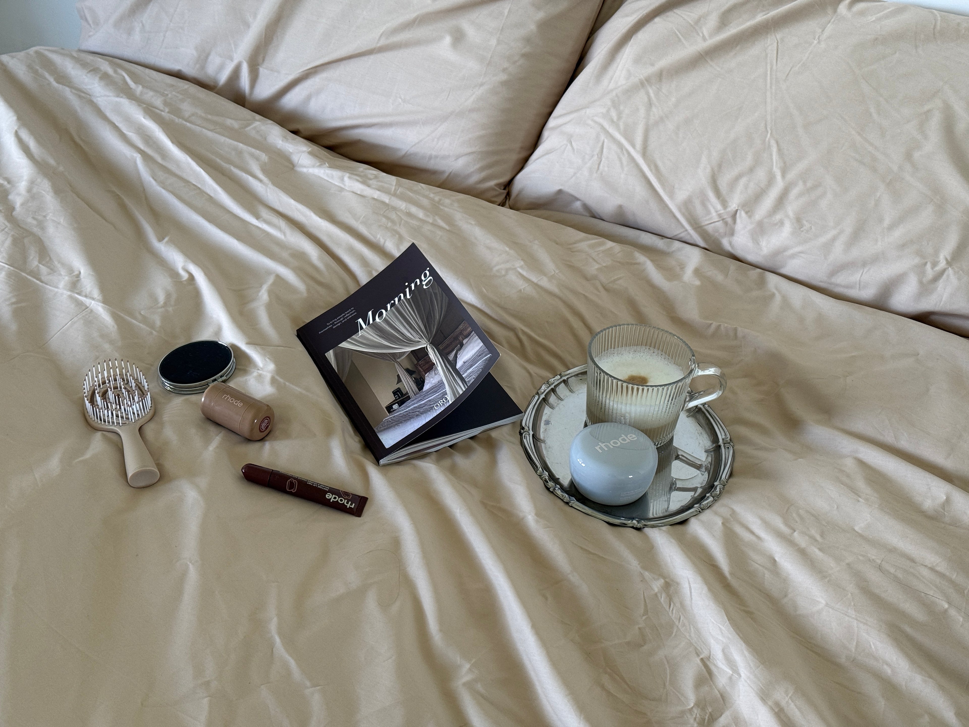 A styled flat-lay on cream-colored bedding featuring a black and white 'Morning' magazine, a white hairbrush, a small black oval compact, a beige hair roller, and a glass mug on a mirrored tray with a white candle.